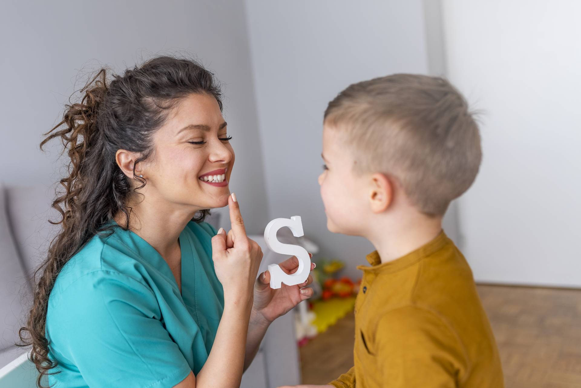 Sprachtherapiesitzung mit einem kleinen Jungen: Logopädin hält den Buchstaben Klotz S in der Hand und zeigt auf ihre Lippen für die Aussprache.