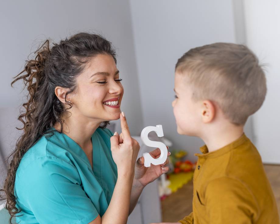 Sprachtherapiesitzung mit einem kleinen Jungen: Logopädin hält den Buchstaben Klotz S in der Hand und zeigt auf ihre Lippen für die Aussprache.