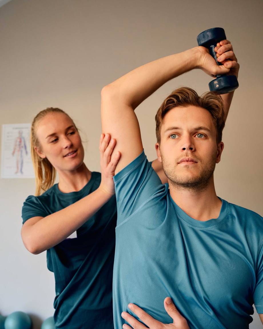 Ein Mann bei der Physiotherapie trainiert mit Hanteln, während eine Frau seine Haltung korrigiert. 