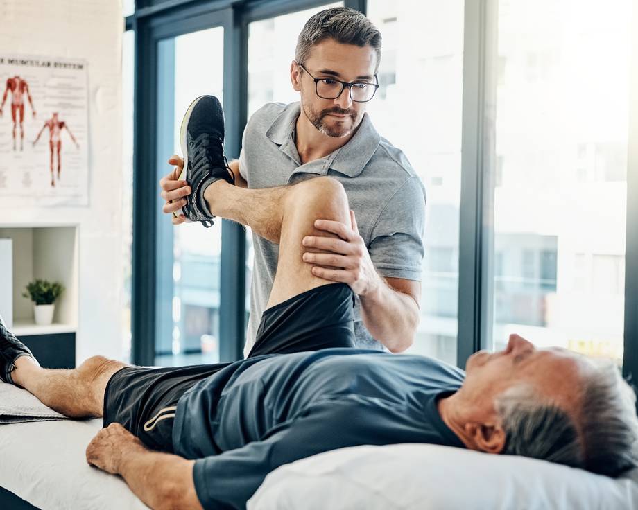 Physiotherapeut beugt Knie von älterem Mann auf einer Liege